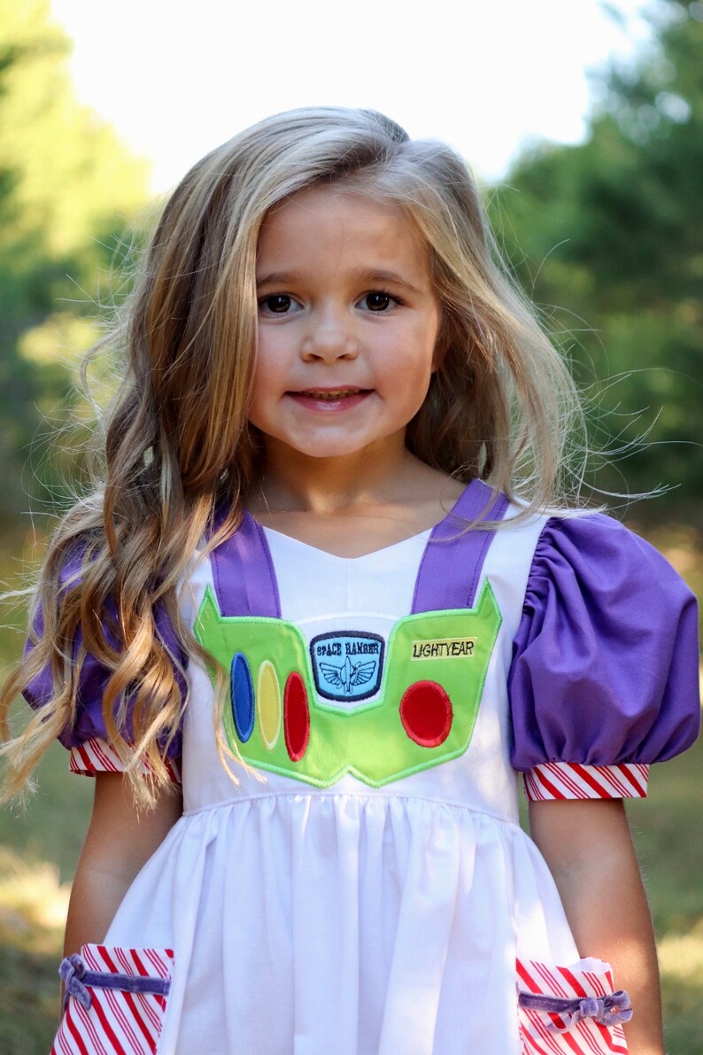 May include: A white dress with purple accents and a green chest piece, resembling a Space Ranger uniform. The dress features the text "LIGHTYEAR" and "SPACE RANGER". The sleeves and cuffs have red and white stripes. The dress is for children.
