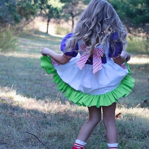 May include: A child's dress with a white bodice, purple sleeves, and a green ruffled skirt. The dress has a red and white striped bow in the back. The child is wearing red high-top sneakers with white laces and striped socks. The background is a grassy area with trees.