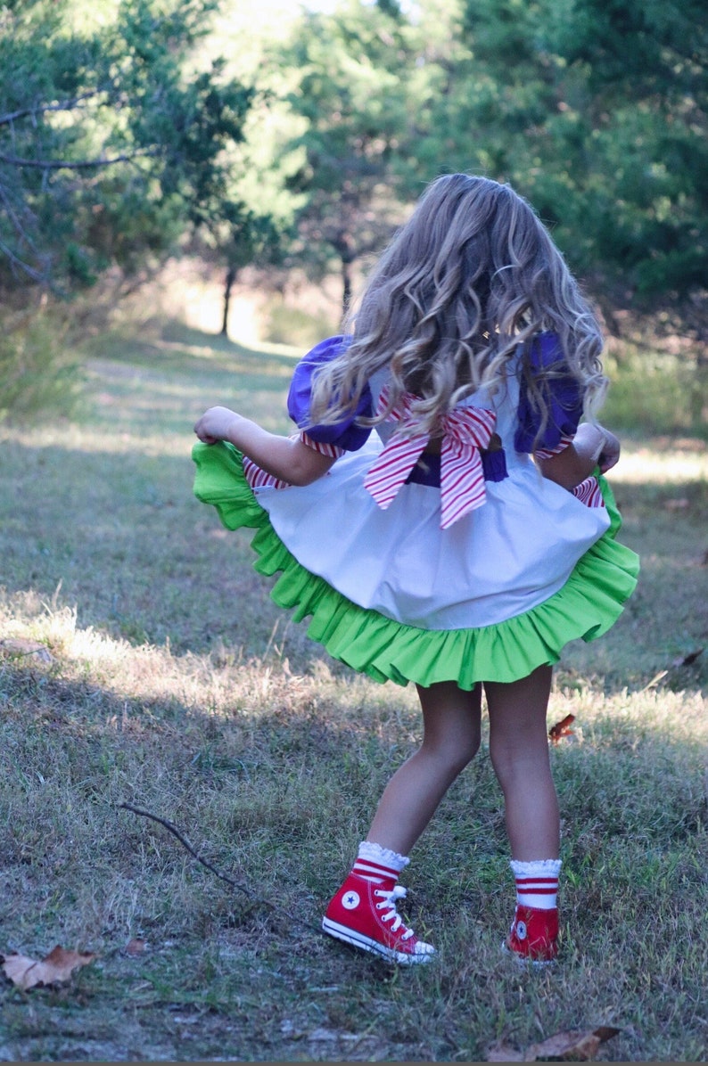 May include: A child's dress with a white bodice, purple sleeves, and a green ruffled skirt. The dress has a red and white striped bow in the back. The child is wearing red high-top sneakers with white laces and striped socks. The background is a grassy area with trees.