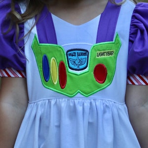 May include: A white dress with purple accents, designed to resemble the character Buzz Lightyear. The dress features a green chest plate with colorful details and the words "SPACE RANGER" and "LIGHTYEAR". The sleeves are purple with red and white striped cuffs.