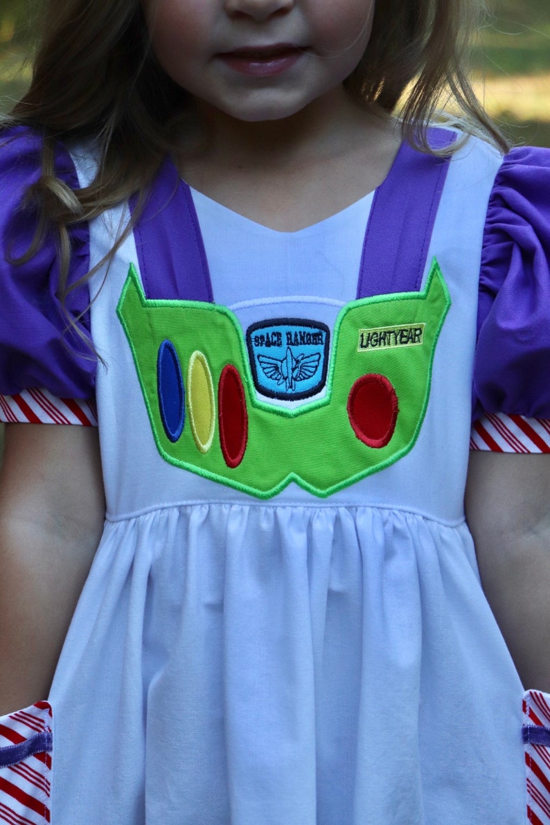 May include: A white dress with purple accents, designed to resemble the character Buzz Lightyear. The dress features a green chest plate with colorful details and the words "SPACE RANGER" and "LIGHTYEAR". The sleeves are purple with red and white striped cuffs.