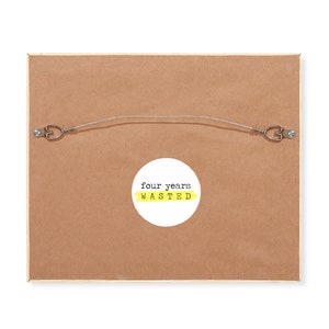 May include: The back of a framed print with a yellow circle sticker that says "four years wasted" in black text.