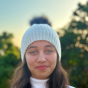 May include: A light blue knit beanie with a dark pom-pom. The ribbed hat is worn by a person with long, dark hair and a white turtleneck sweater. The background is a soft focus of green trees and a blue sky.