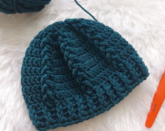 Op de afbeelding: Een tealblauwe gebreide beanie met een textuurpatroon. De beanie ligt op een wit, pluche oppervlak.
