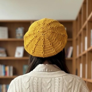 May include: A mustard yellow crochet beret featuring a radial pattern of textured stitches and openwork details. The hat has a circular crown with a structured, slightly raised design that radiates from the centre towards the outer edge.