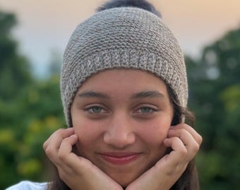 May include: A light gray knit beanie with a dark gray pom-pom. The beanie is worn by a person with long dark hair and a white shirt. The person is smiling and resting their chin on their hands.