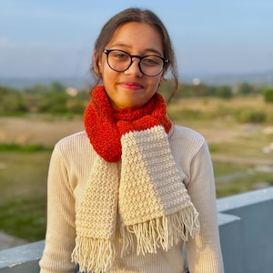May include: A handmade scarf with a two-tone design. The top section is a vibrant red, transitioning to a cream-colored lower section with fringe. The scarf appears to be knitted or crocheted, offering a cozy and textured look. The scarf is worn around the neck.