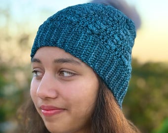 May include: A teal-colored knitted beanie with a textured pattern and a gray pom-pom. The hat is made of a soft yarn and is designed to keep the head warm. The beanie is a stylish accessory for cold weather.