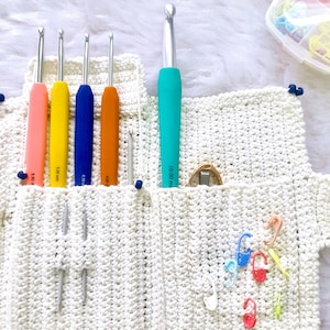 Puede incluir: Una bolsa blanca de ganchillo con asa, que contiene una variedad de agujas de ganchillo de diferentes colores y tamaños, incluyendo una aguja de 5,50 mm, 5,00 mm, 4,50 mm, 4,00 mm y una aguja de 10,00 mm. La bolsa también contiene una pequeña herramienta de metal y una variedad de marcadores de punto de colores.