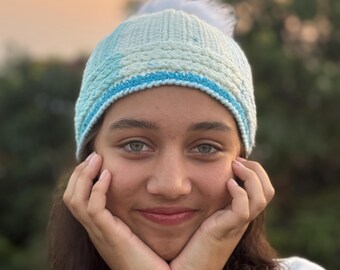 Könnte beinhalten: Eine hellblaue und weiße Strickmütze mit einem flauschigen weißen Bommel. Die Mütze hat ein blaues Band und ein strukturiertes Muster. Die Person trägt ein weißes Hemd und hat lange dunkle Haare.