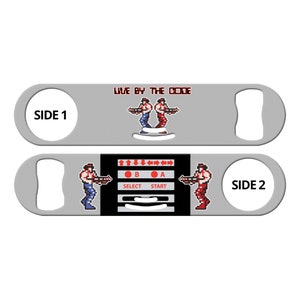May include: A silver bottle opener with a black and white video game design. The design features two pixelated characters holding guns and a control panel with buttons labeled "A", "B", "Select", and "Start". The text "Live by the Code" is printed on the top of the bottle opener. The words "Side 1" and "Side 2" are printed on the sides of the bottle opener.