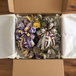 May include: A football-themed wreath with purple, gold, and brown ribbons, football accents, and a yellow helmet. The wreath is inside a cardboard box with white tissue paper.