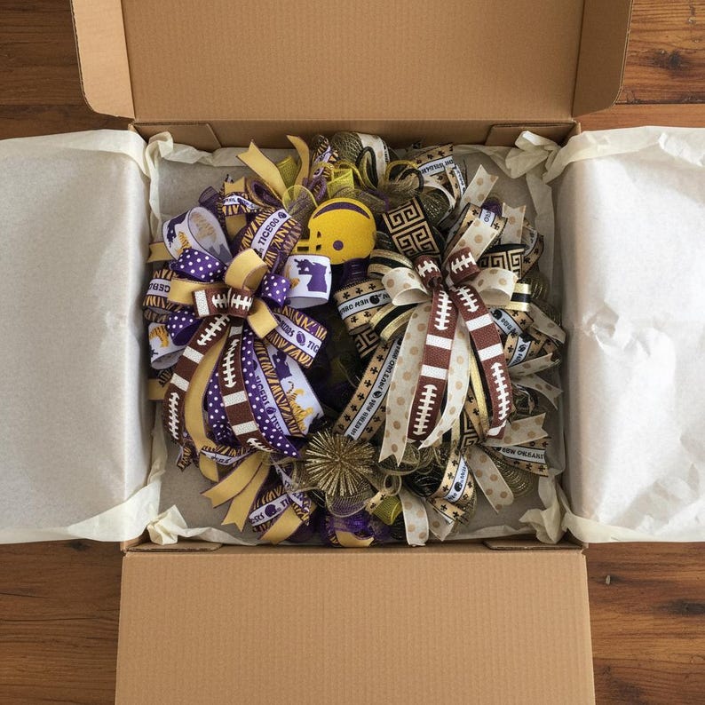 May include: A football-themed wreath with purple, gold, and brown ribbons, football accents, and a yellow helmet. The wreath is inside a cardboard box with white tissue paper.