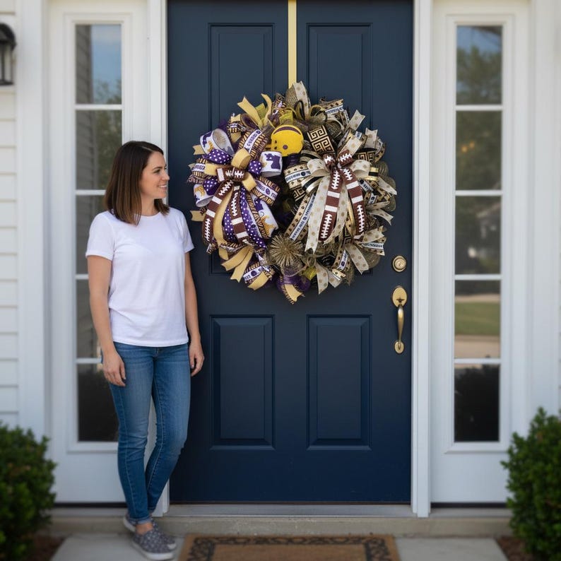 May include: A decorative wreath with football-themed ribbons and accents hangs on a navy blue door. The wreath features ribbons in shades of brown, gold, purple, and white, with football and team logo patterns. A yellow football-shaped ornament is centered in the wreath.