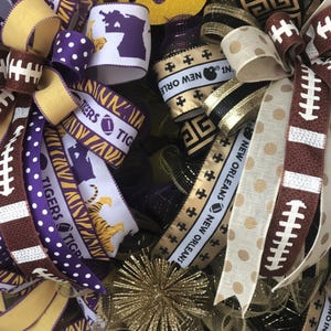 May include: A decorative wreath with a mix of ribbons in purple, gold, and brown. Some ribbons feature footballs, polka dots, and the text "Tigers" and "New Orleans". A gold starburst ornament is also visible.