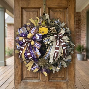 May include: A decorative wreath with football and team-themed ribbons in purple, gold, and brown. The wreath includes a miniature yellow football helmet and decorative accents, perfect for sports fans. The wreath is hanging on a wooden door.