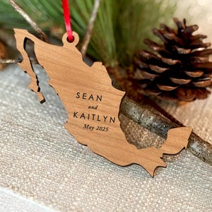 May include: A wooden Mexico-shaped ornament, engraved with "Sean and Kaitlyn" and "May 2025". It has a red ribbon for hanging. The ornament is displayed with pine needles and a pine cone, creating a festive look.