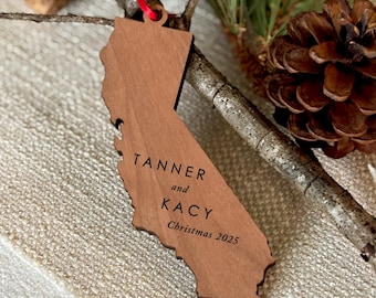 May include: A wooden California-shaped ornament, engraved with "Tanner and Kacy" and "Christmas 2025". It hangs by a red ribbon, with pine needles and a pine cone in the background. A festive, personalised decoration.