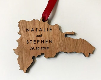May include: Wooden ornament shaped like the Dominican Republic, engraved with "NATALIE and STEPHEN" and the date "10.20.2018". A red ribbon is attached for hanging. The ornament is made of light brown wood.