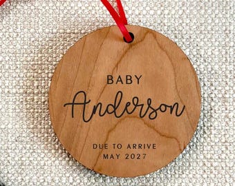 May include: A round wooden ornament with a red ribbon for hanging. The text reads BABY Anderson DUE TO ARRIVE MAY 2027. The wood has a natural light brown grain pattern, and the lettering is printed in a dark, clear font across the center surface.