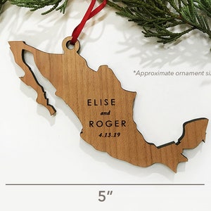 May include: A wooden Mexico-shaped ornament, approx. 12.7 cm long and 8.9 cm wide. It features the engraved names "ELISE and ROGER" and the date "4.13.19". The ornament is suspended by a red ribbon, suitable for hanging.