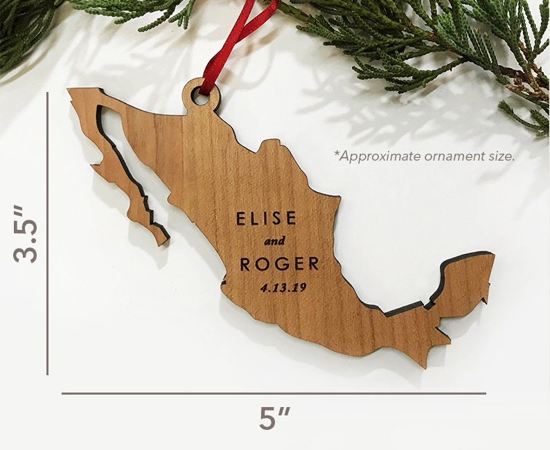 May include: A wooden Mexico-shaped ornament, approx. 12.7 cm long and 8.9 cm wide. It features the engraved names "ELISE and ROGER" and the date "4.13.19". The ornament is suspended by a red ribbon, suitable for hanging.