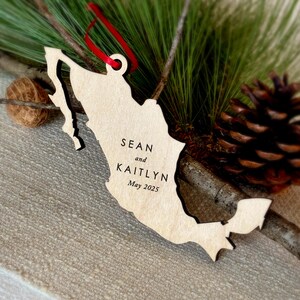 May include: A wooden Mexico-shaped ornament with the names "Sean and Kaitlyn" and the date "May 2025" engraved. It has a red ribbon for hanging, displayed with pine needles and a pine cone, creating a festive feel.