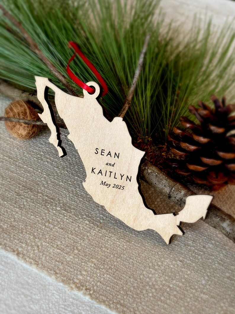 May include: A wooden Mexico-shaped ornament with the names "Sean and Kaitlyn" and the date "May 2025" engraved. It has a red ribbon for hanging, displayed with pine needles and a pine cone, creating a festive feel.