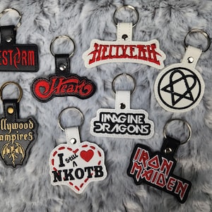May include: Nine different keychains with various band names and logos. The keychains are made of black and white leather with red embroidery. The band names include Halestorm, Heart, Hollywood Vampires, Imagine Dragons, Iron Maiden, NKOTB, and a pentagram.