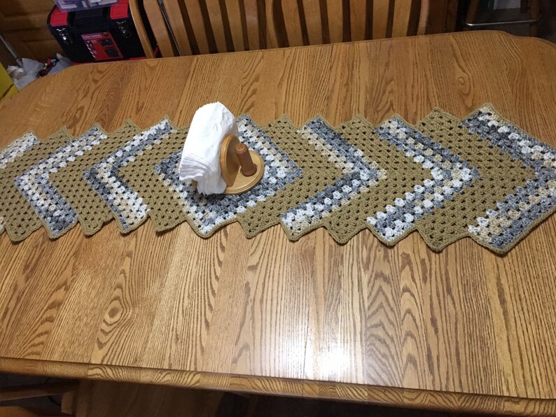 Puede incluir: Camino de mesa de ganchillo hecho a mano con un patr&oacute;n de diamantes, en tonos beige, gris y blanco. El camino de mesa est&aacute; sobre una mesa de madera, con un servilletero de madera y servilletas blancas en el centro.