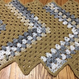 Puede incluir: Camino de mesa de ganchillo hecho a mano con borde en zigzag. El camino de mesa presenta un patr&oacute;n repetitivo de cuadrados de la abuela en tonos beige, gris y blanco. El trabajo de ganchillo es visible, con una apariencia texturizada. El camino de mesa est&aacute; colocado sobre una superficie de madera.