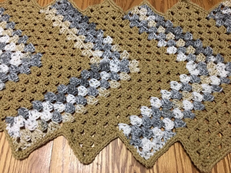 Puede incluir: Camino de mesa de ganchillo hecho a mano con borde en zigzag. El camino de mesa presenta un patr&oacute;n repetitivo de cuadrados de la abuela en tonos beige, gris y blanco. El trabajo de ganchillo es visible, con una apariencia texturizada. El camino de mesa est&aacute; colocado sobre una superficie de madera.