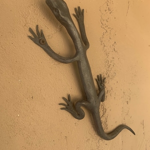 May include: A black metal lizard sculpture mounted on a tan wall. The lizard has a long tail and detailed features.