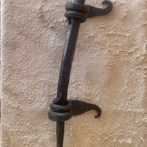 Op de afbeelding: Een zwarte metalen deurgreep met een decoratieve draai en een haak aan de zijkant.