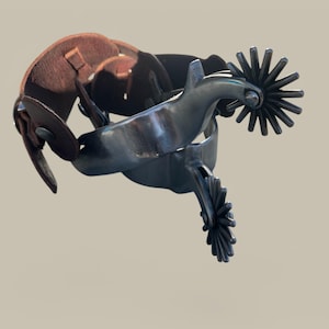 May include: A pair of silver spurs with a brown leather strap. The spurs have a star-shaped design and are attached to a leather strap.