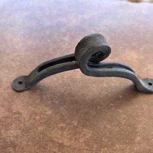 May include: Black metal door handle with a decorative scroll design. The handle has two mounting holes on the bottom.