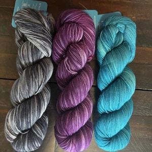 May include: Three skeins of yarn in shades of gray, purple, and teal. The yarn is twisted into a thick, textured bundle. Each skein has a tag attached. The yarn is displayed on a dark wooden surface.