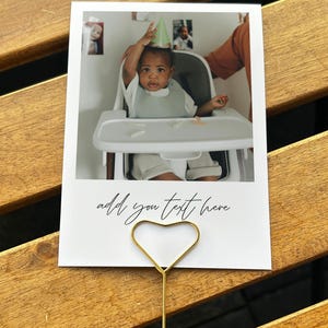 May include: A white photo card with a gold heart-shaped holder. The photo shows a baby in a high chair wearing a party hat. The text "add you text here" is written on the card.