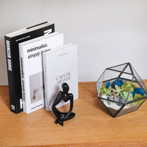K&ouml;nnte beinhalten: Drei B&uuml;cher mit den Titeln Moncur Home, Minimalist Simply Living und Caf&eacute; du Monde, eine schwarze abstrakte Denker-Figur und ein geometrisches Glasterrarium mit gr&uuml;nen Sukkulenten, blauem Moos und wei&szlig;en Kieselsteinen auf Holz.