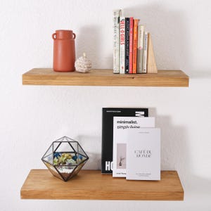K&ouml;nnte beinhalten: Zwei horizontale, hellbraune Holzregale, die an einer wei&szlig;en Wand montiert sind. Das obere Regal enth&auml;lt eine Terrakotta-Vase, eine kleine geflochtene Kugel und mehrere B&uuml;cher. Das untere Regal zeigt ein geometrisches Glas-Terrarium.