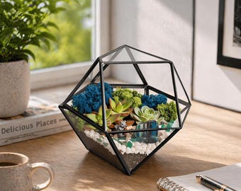 May include: A geometric glass terrarium with a black metal frame containing green and blue preserved moss, small succulent plants, white pebbles, and a miniature gray animal figurine. The container has a multi-faceted, diamond-like shape.