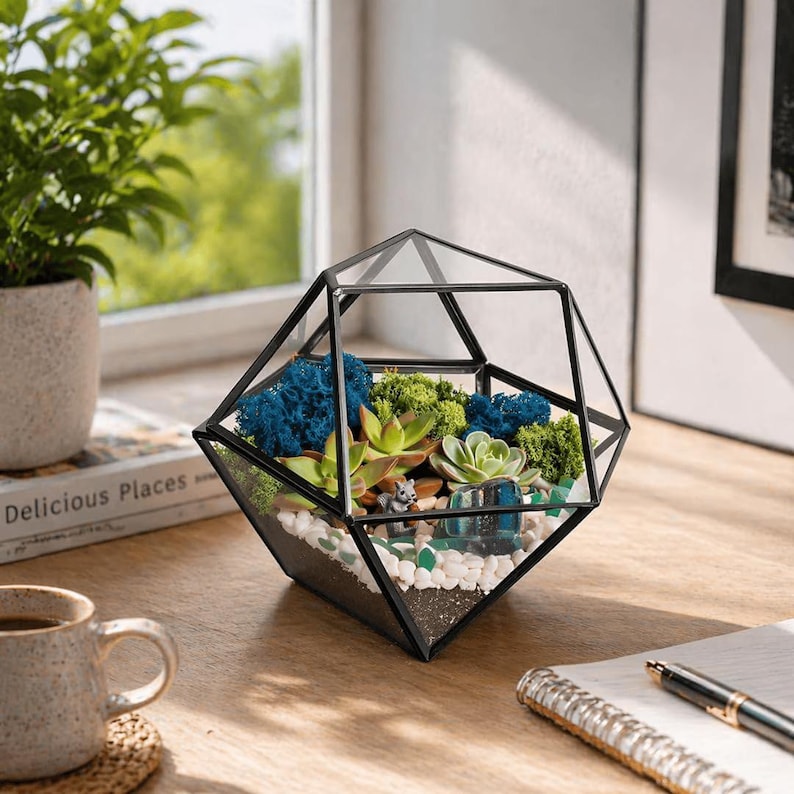 K&ouml;nnte beinhalten: Ein geometrisches Glasterrarium mit schwarzem Metallrahmen, gef&uuml;llt mit gr&uuml;nem und blauem konserviertem Moos, kleinen Sukkulenten, wei&szlig;en Kieselsteinen und einer grauen Miniatur-Tierfigur. Das Gef&auml;&szlig; hat eine facettierte Diamantform.