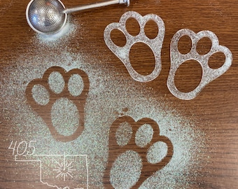Op de afbeelding: Vier transparante plastic koekjesvormen in de vorm van een poot afdruk op een houten oppervlak bedekt met witte glitter. De koekjesvormen zijn in verschillende maten en vormen. De tekst "405 Crafted" is zichtbaar in de linkerbenedenhoek van de afbeelding.