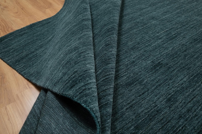 May include: A folded teal rug with a textured surface. The rug's fibers create a subtle diagonal pattern. The rug is displayed on a wooden floor, with a portion of the rug folded over, revealing its underside.