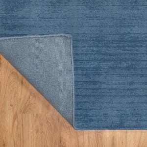 May include: A blue rug with a textured, horizontal striped pattern. The corner of the rug is folded back, revealing a lighter blue backing. The rug is placed on a light brown wooden floor.