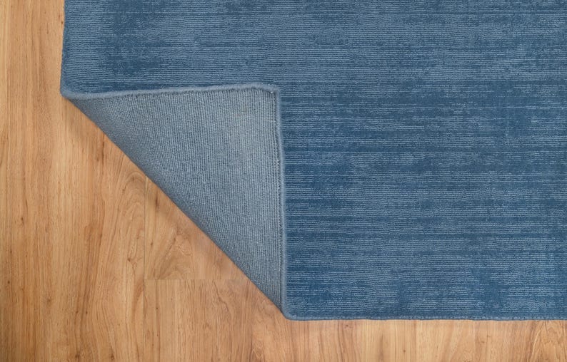 May include: A blue rug with a textured, horizontal striped pattern. The corner of the rug is folded back, revealing a lighter blue backing. The rug is placed on a light brown wooden floor.