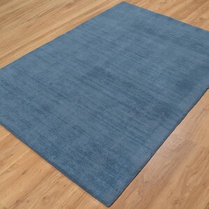 May include: A rectangular, blue rug with a textured surface. The rug is placed on a wooden floor with a light brown color. The rug's color is a solid, medium shade of blue.