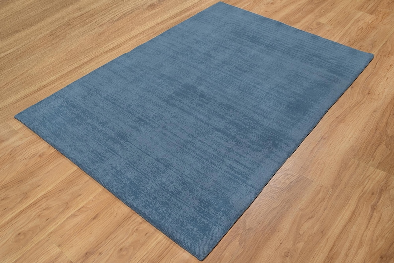 May include: A rectangular, blue rug with a textured surface. The rug is placed on a wooden floor with a light brown color. The rug's color is a solid, medium shade of blue.