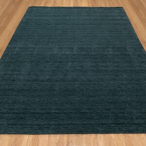 May include: A large, rectangular area rug in a solid teal color. The rug has a textured appearance with horizontal lines, and it is placed on a light brown wooden floor. The rug's color is consistent across its surface, providing a simple, modern aesthetic.