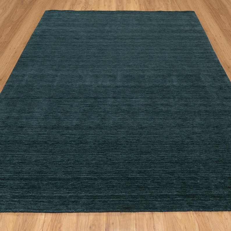 May include: A large, rectangular area rug in a solid teal color. The rug has a textured appearance with horizontal lines, and it is placed on a light brown wooden floor. The rug's color is consistent across its surface, providing a simple, modern aesthetic.